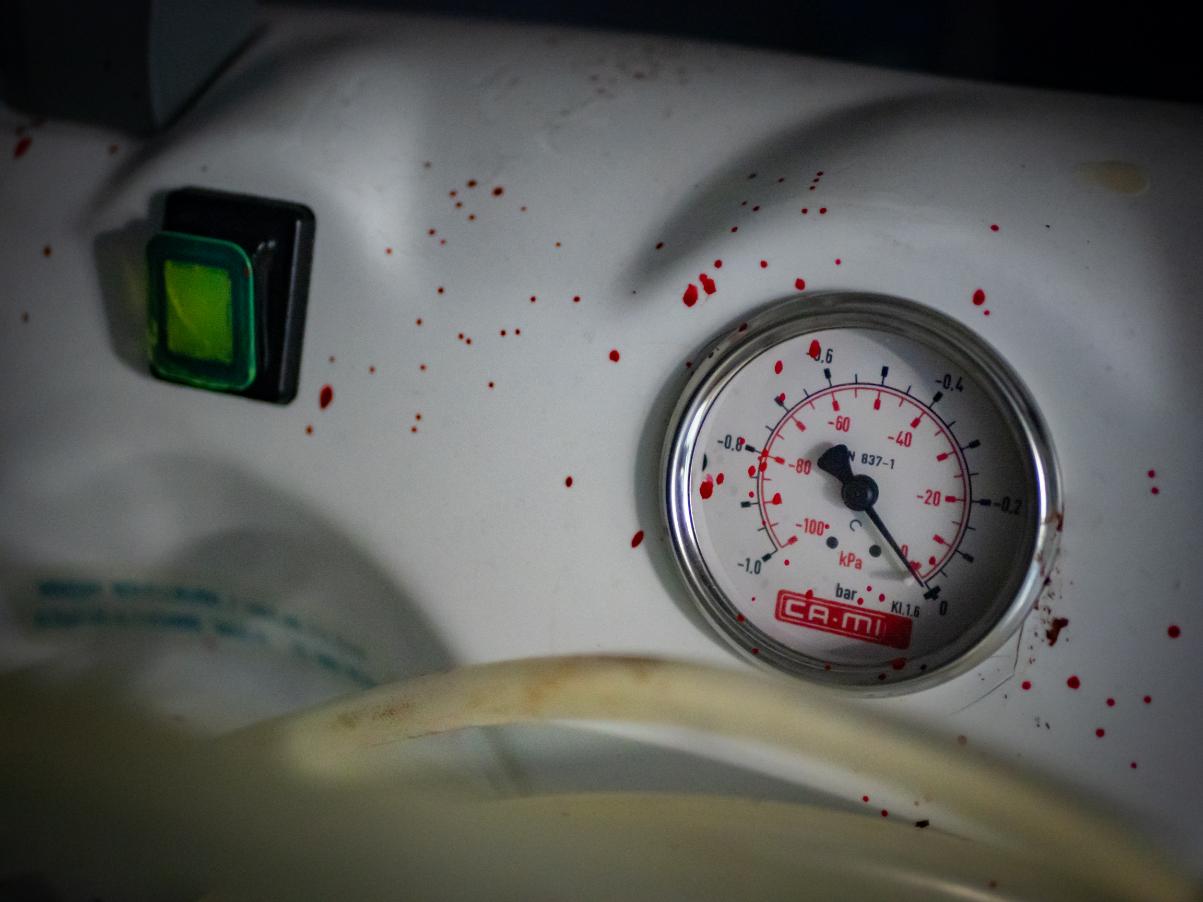 Primer plano de un respirador médico con un manómetro y un botón verde iluminado, salpicado de gotas de sangre simulada. Atrezzo inmersivo de terror en Clínica El Pasaje Escape Room en A Coruña.