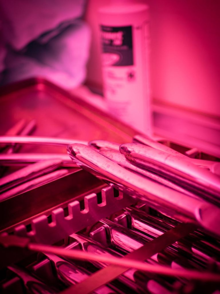 Primer plano de una bandeja metálica con instrumental quirúrgico, incluyendo dilatadores médicos, bajo una dramática luz magenta en Clínica El Pasaje Escape Room en A Coruña.