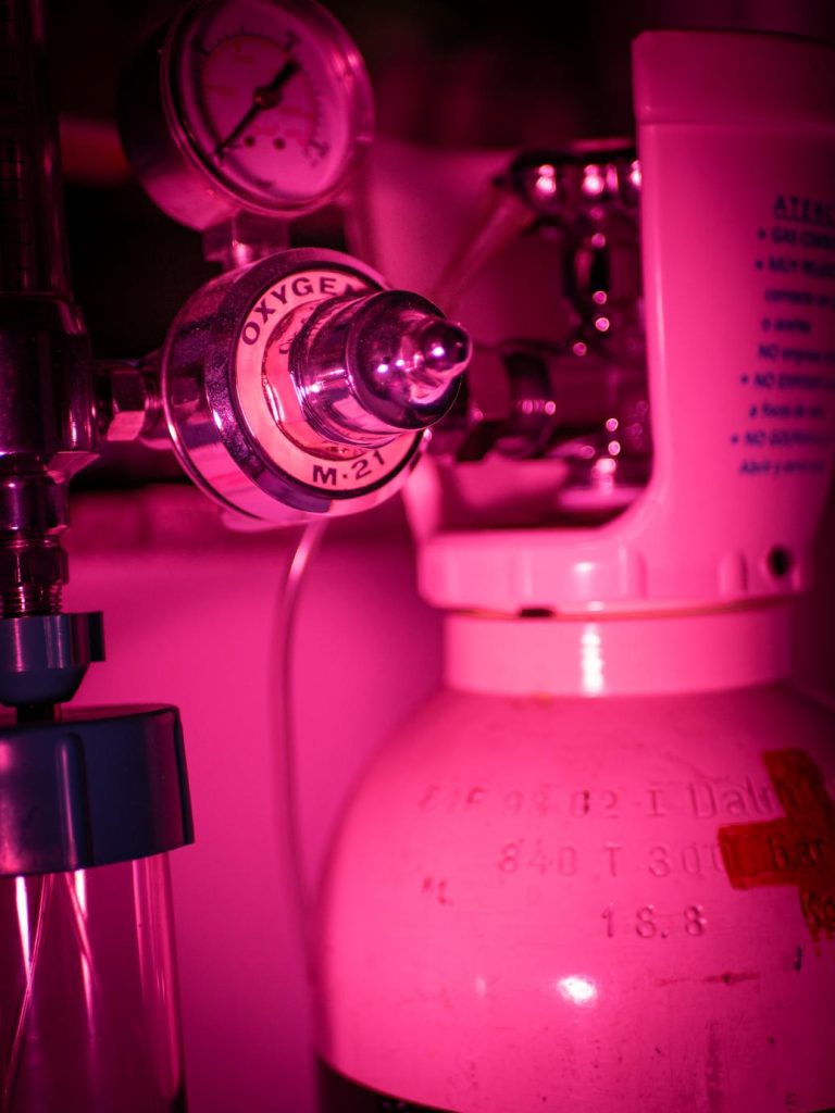 Primer plano de una bombona de oxígeno médico con regulador y manómetro bajo una inquietante luz magenta, elemento de ambientación en Clínica El Pasaje Escape Room en A Coruña.