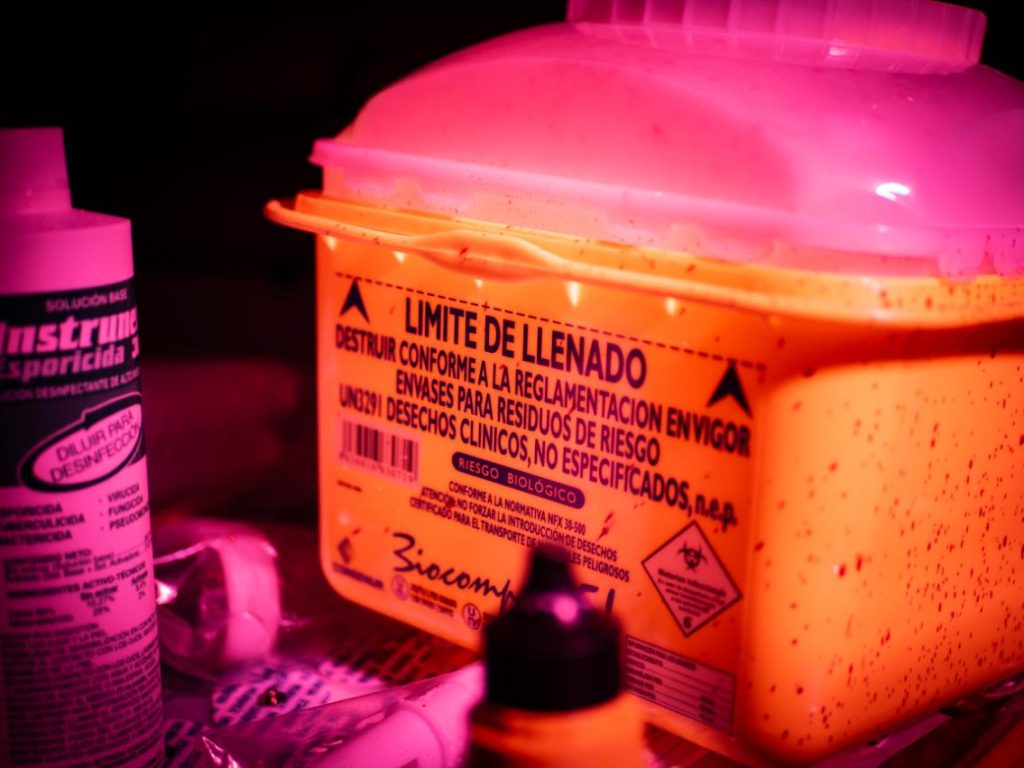 Primer plano de un contenedor amarillo para residuos biológicos y líquidos desinfectantes bajo una tensa luz roja, parte del atrezzo de material médico en Clínica El Pasaje Escape Room en A Coruña.