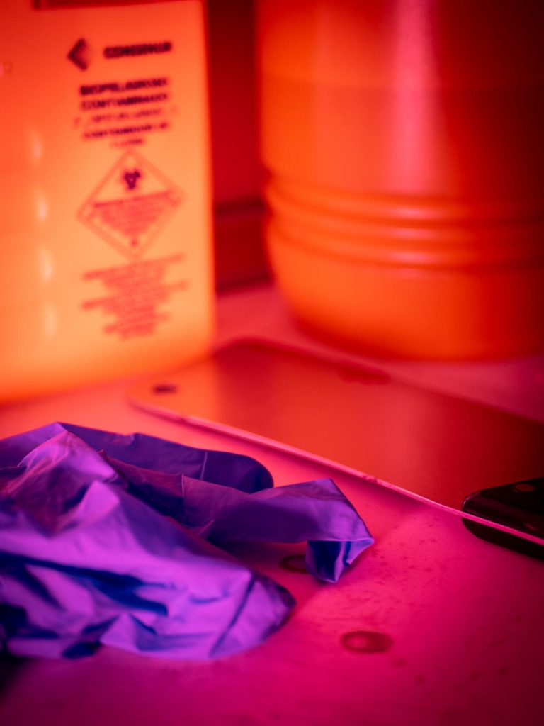 Primer plano bajo una inquietante luz roja mostrando un contenedor de riesgo biológico, un cuchillo metálico de gran tamaño y guantes médicos de nitrilo morados. Detalle de la ambientación macabra e inmersiva en Clínica El Pasaje Escape Room en A Coruña.