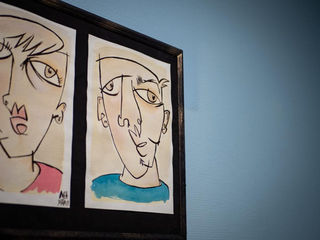 Cuadro decorativo con ilustraciones abstractas e inquietantes de rostros colgado en una pared azul, parte de la ambientación de Clínica El Pasaje Escape Room en A Coruña.