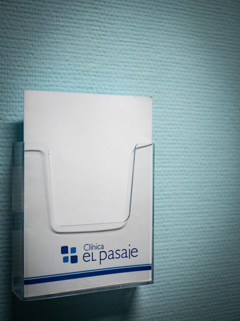 Portafolletos de pared de acrílico transparente que contiene papel con el logotipo de Clínica El Pasaje Escape Room en A Coruña sobre una pared azul texturizada.