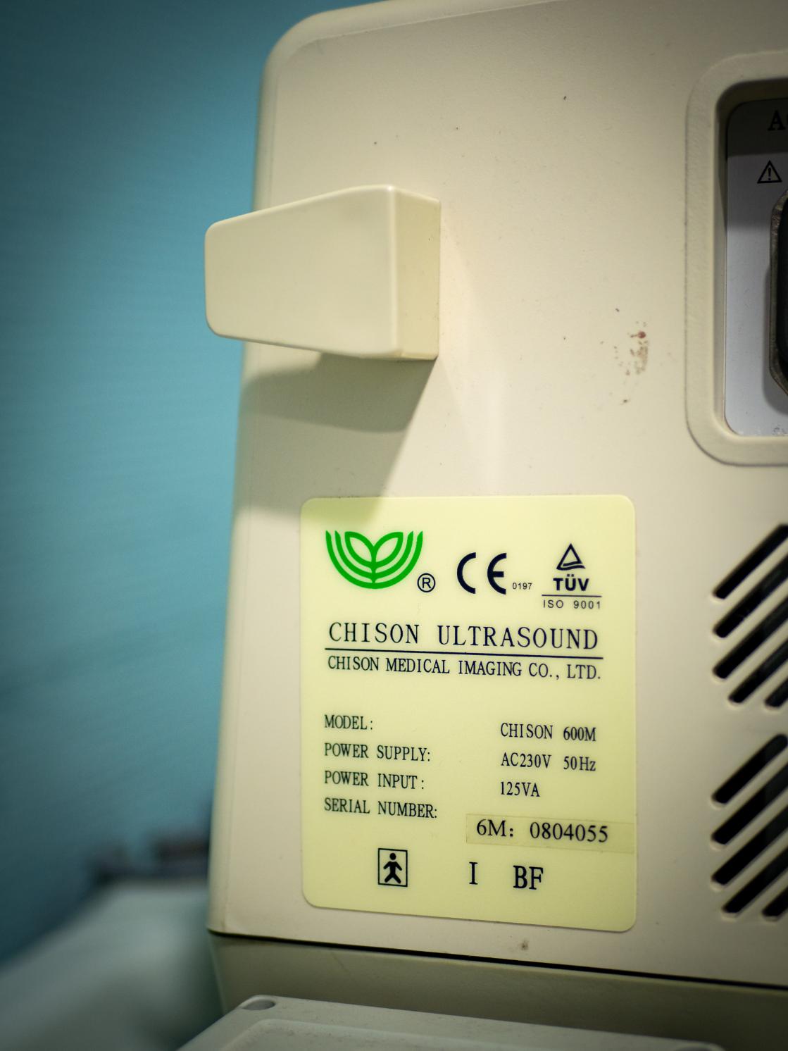 Primer plano detallado de un ecógrafo médico vintage marca ATL encendido, mostrando una imagen borrosa, en un entorno de escape room oscuro con paredes de piedra rústica y tenue iluminación. La máquina tiene perillas y un teclado. Una sonda descansa al lado.