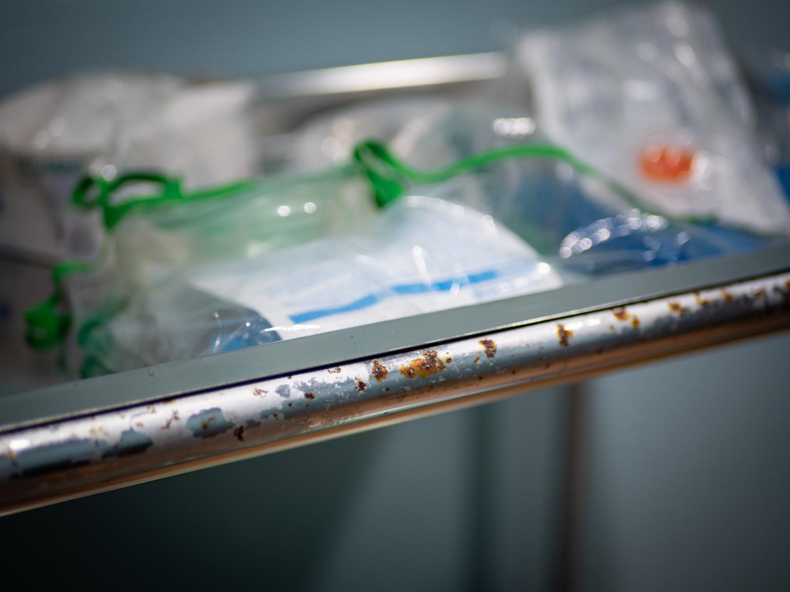 Detalle elevado y en ángulo de un antiguo carrito médico metálico de varios niveles en Clínica El Pasaje Escape Room, A Coruña. Los estantes están repletos de botellas de boticario de vidrio, viales, instrumental quirúrgico vintage y vendajes, con etiquetas envejecidas y un candado visible